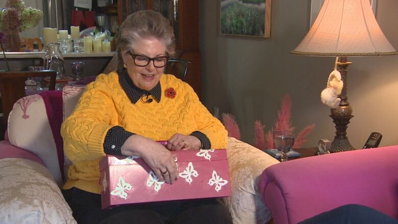 Woman with brown hair and glasses, wearing a yellow sweater, sits on a pink and white armchair, and holds a pink metal toolbox on her lap.