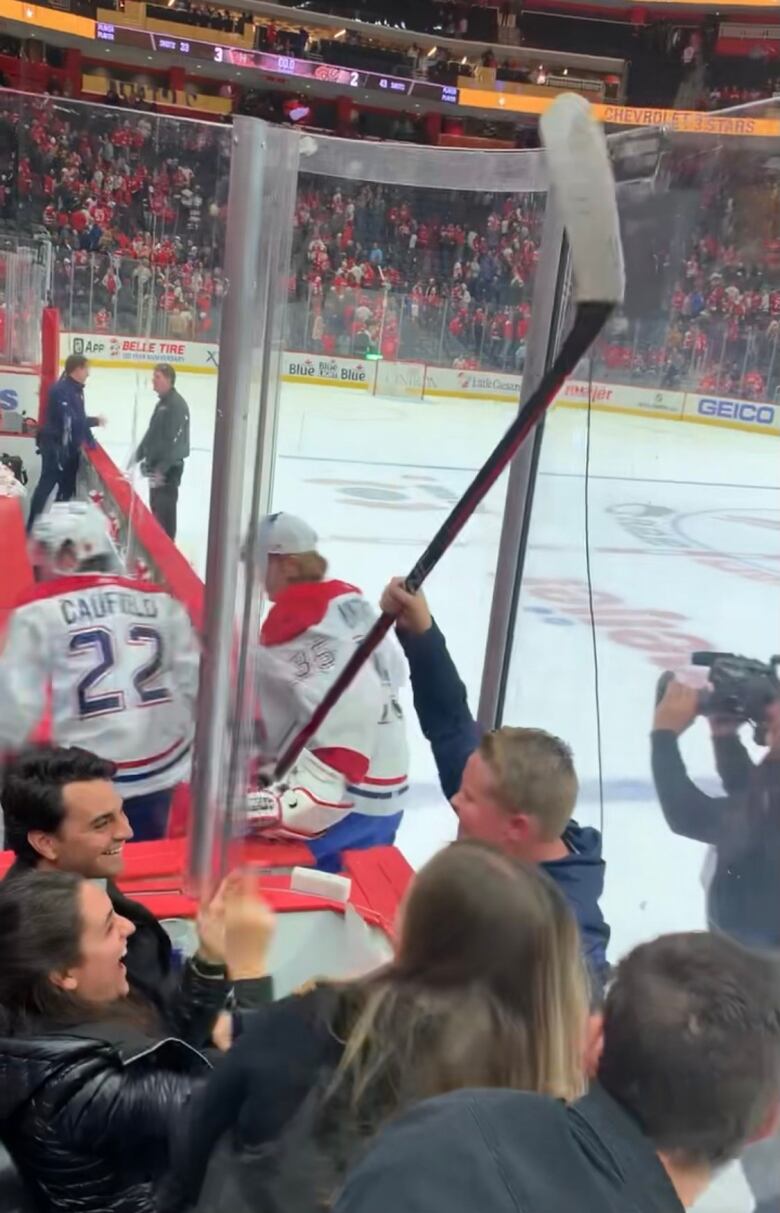 A boy in the stands of a hockey arena holds up a hockey stick as the crowd around him smiles. The backs of the players can be seen on the opposite side of the boards. 