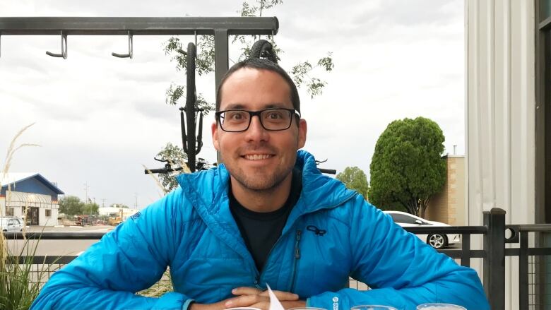 A white man with glasses sits at a desk. He is smiling, facing the camera. He is wearing a blue sweater.