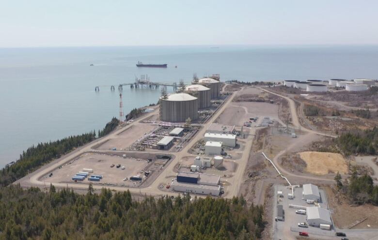 An aerial view of an industrial facility along a shoreline, featuring three large metal cylinders used for storage of liquefied natural gas. 