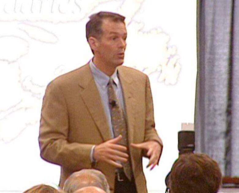 A middle-aged man with a beige blazer speaks in front of a meeting. A projection screen behind him shows an outline of parts of the Atlantic provinces. 