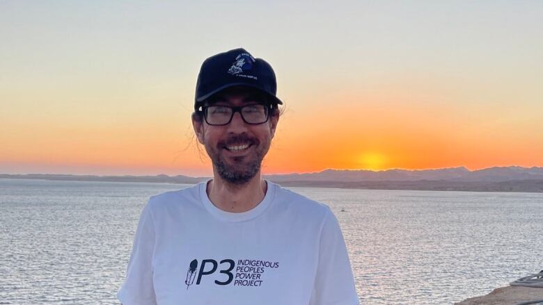 A man smiles and wears a baseball cap and sunglasses, as he stands in front of the ocean at sunset.