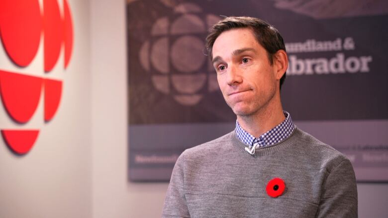 A man sits in a chair with a grey sweater overtop of a blue button up. He is wearing a poppy on his left side. 