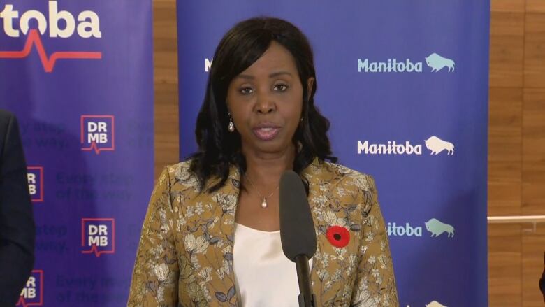 A woman wearing a floral blazer with a red poppy on the lapel speaks into a microphone at a podium.