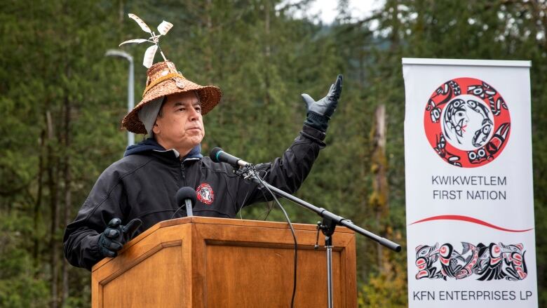 A man raises his left hand as he gives a talk at a podium, in the outdoors.