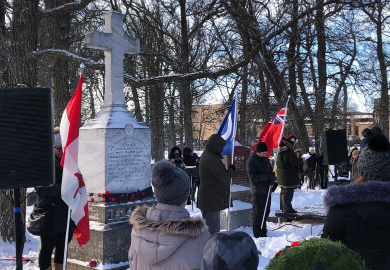 People holding flags stand around a tall stone marker topped with a cross, while several others stand in the snow around them.