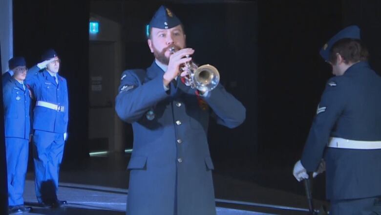 A man in a navy blue military uniform stands in a spotlight on a darkened stage, playing a bugle, as other uniformed people stand behind him.