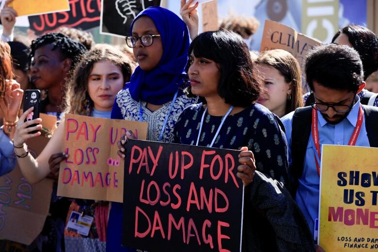 A young woman with shoulder-length brown hair and a nose ring stands at the front of a crowd of protesters, holding a sign that reads: 