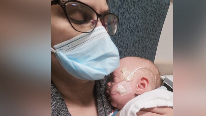 A woman wearing a mask cradles a baby in her arms. A tube is taped to side of the baby's face.