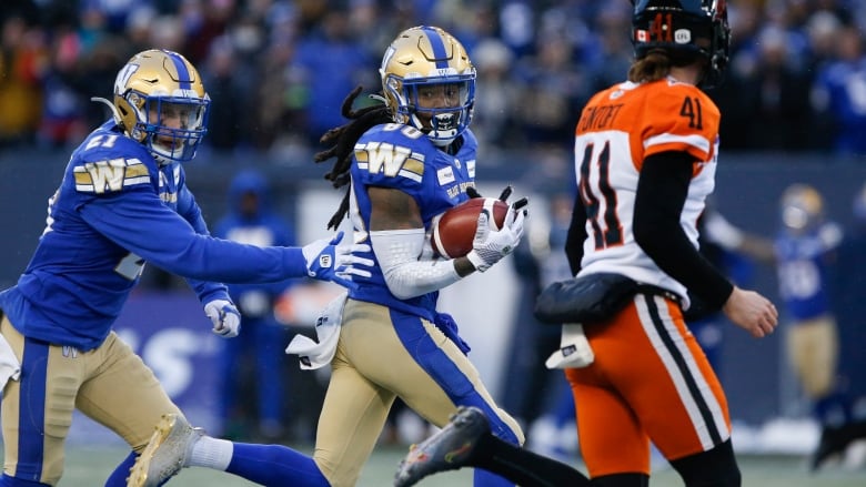 A player wearing a blue jersey runs next to a player wearing an orange and white jersey while carrying a football.