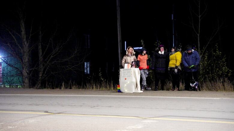 A table on the side of the road has a white table cloth, photographs and the names of people who have died. Five people stand behind it along a busy road. 