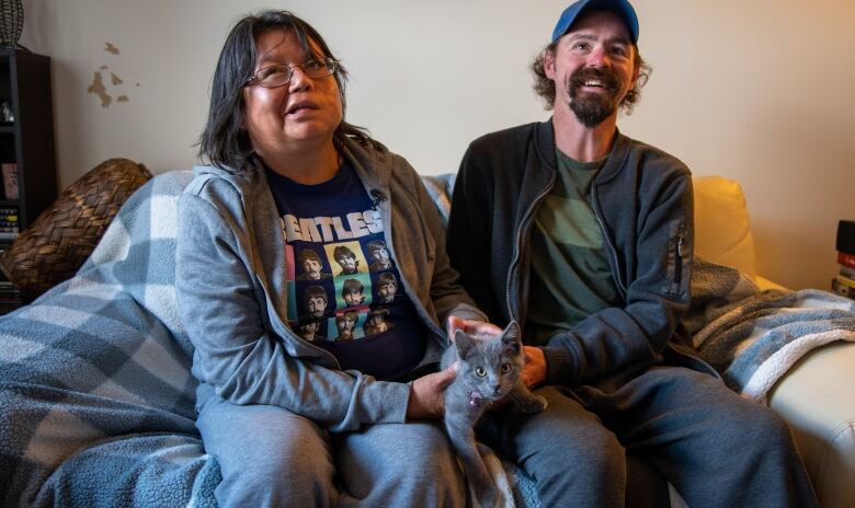 A man and a woman sit on the couch with their cat and smile at the camera.