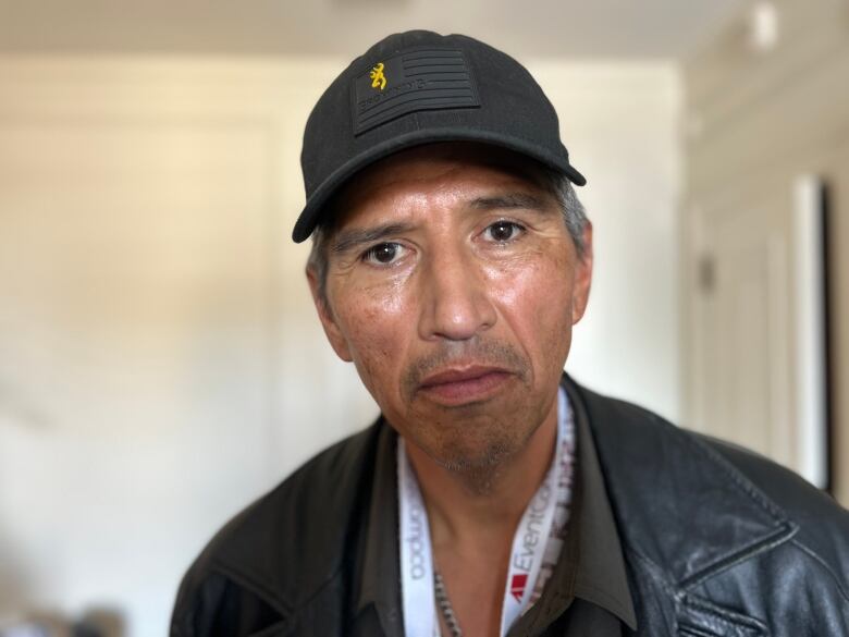 A man wearing a black baseball hat and leather jacket is looking directly at the camera.