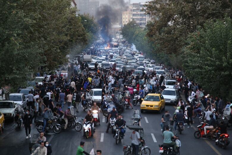 Protesters and vehicles crowd a street as a fire burns in the background.