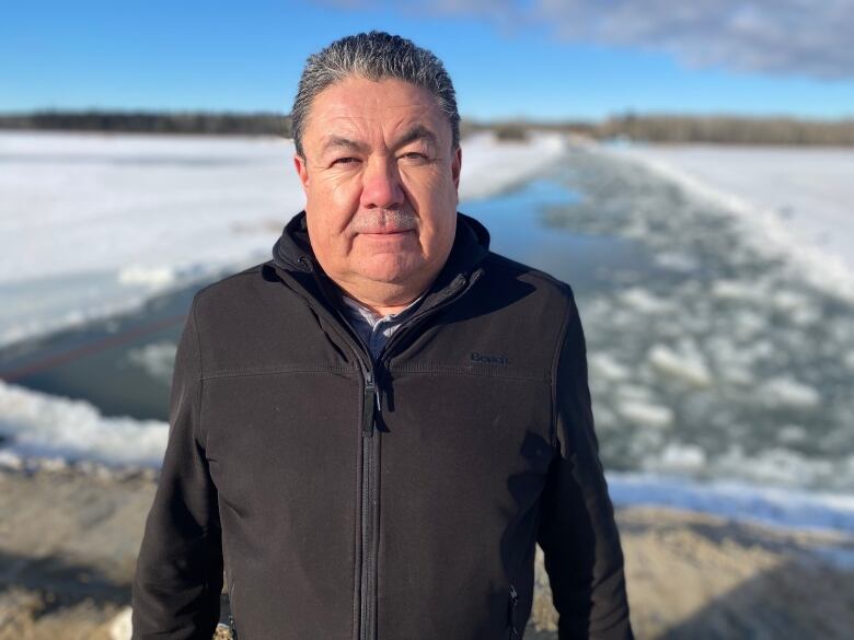 Wearing a black jacket, Wayne Anderson stands facing the camera in front of the Nelson River, which is mostly covered in snow and ice.