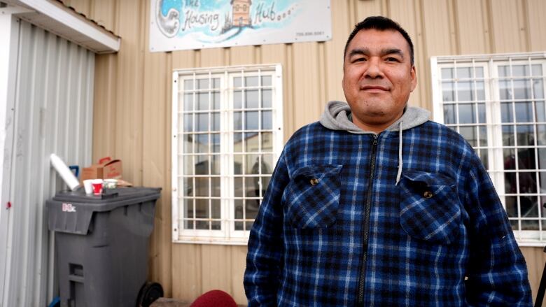 A man in a blue plaid jacket stands in front of a tan building. The building has trash out in front and a sign reading 'The Housing Hub.' 