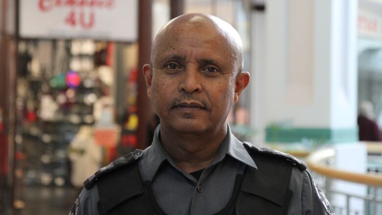 A person wearing a security uniform looks at the camera. A store is in the background.