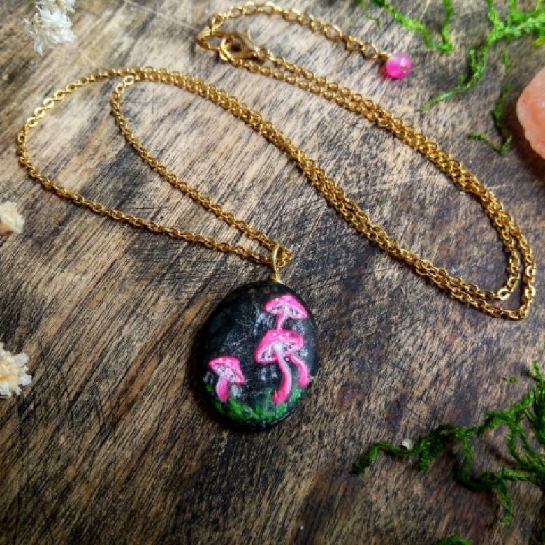 A black medallion with bright pink mushrooms attached to a gold chain is displayed against a wood backdrop