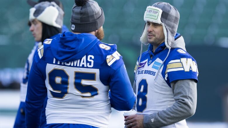 A man wearing a football jersey and a cap with ear flaps stands on a football field, talking to another man in a jersey with his back to the camera.