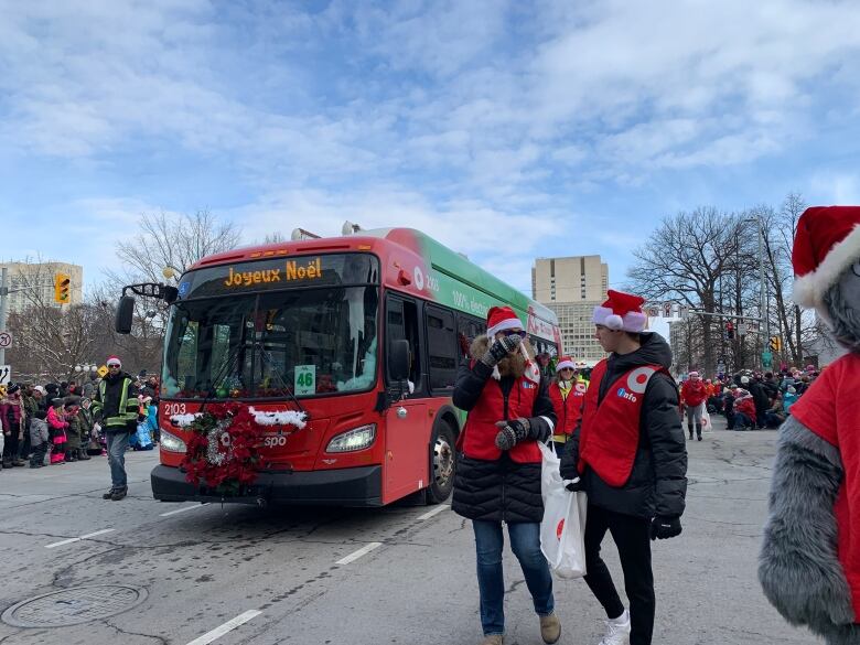 A city bus makes its way along a Christmas parade route while people in Santa Claus hats walk beside it.