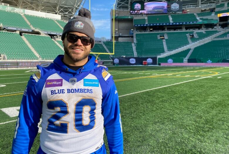 A man in a toque, sunglasses, and blue and white jersey stands in an empty football field smiling.