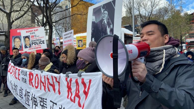 a group of protesters hold a banner saying Justice for Ronny Kay and holding posters, a man speaks into a megaphone