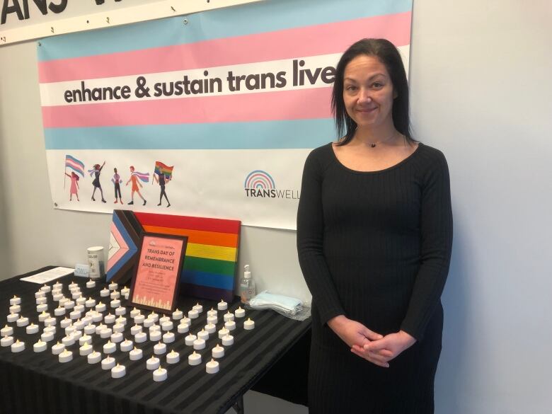 A woman wearing a black dress standing next to a desk which has electric candles and a Pride flag on it and standing in front of the Transgender flag