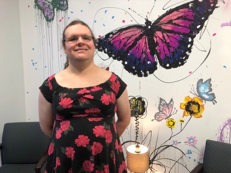 A transgender woman wearing glasses and a flowery dress standing in front of a wall with butterflies painted on it