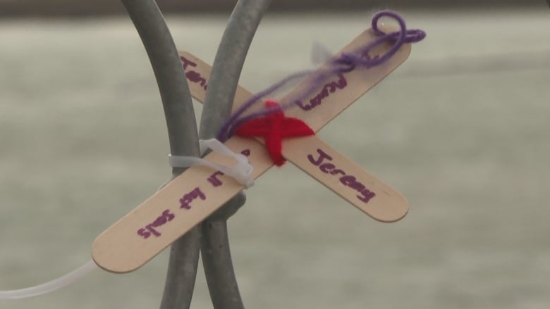 A small wooden cross with the name Jeremy written on it is attached to a metal bridge railing. 