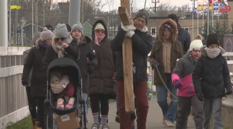 A group of 20 people in winter clothes walk toward the camera, led by a man carrying a large wooden cross