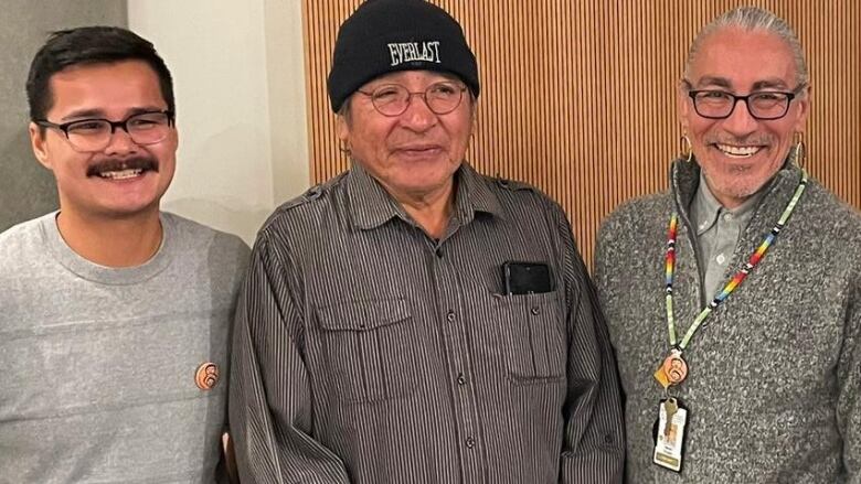 Three Indigenous people stand shoulder-to-shoulder, posing for a picture. They are all smiling and wearing glasses.