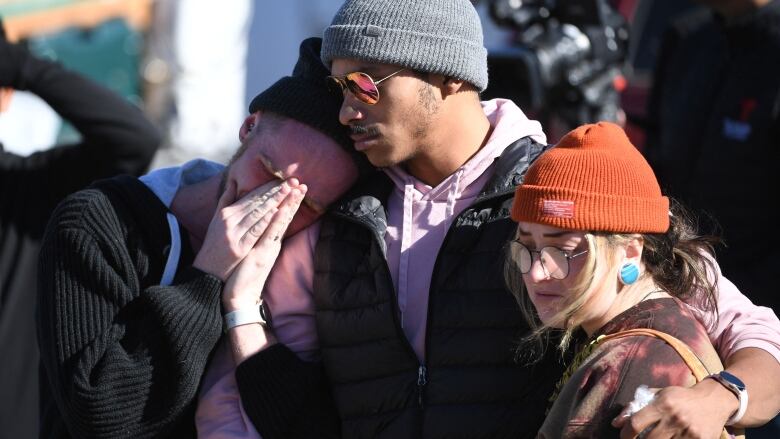 Three people, dressed in winter clothing, stand side by side, their arms around each other. Two are frowning and the other is crying with their hands folded in front of their face. 