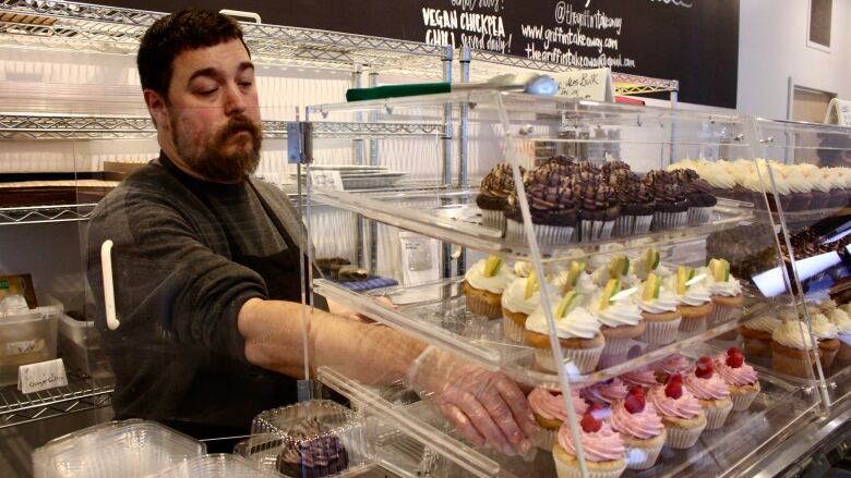 A man wearing an apron stands behind a counter in a bakery, reaching into a display case filled with different flavours of cupcakes. 