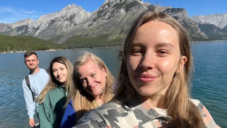 A group of four people look into the camera for a selfie standing in front of a mountain lake. 