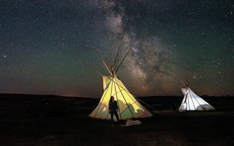 A photo of a young woman standing in front of a teepee in Southern Alberta under a night sky.