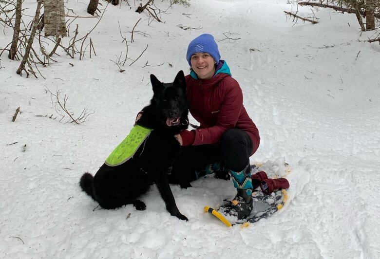 A girl and her dog sit and smile on a snowy forest trail. The girl has a pair of snowshoes sitting next to her.