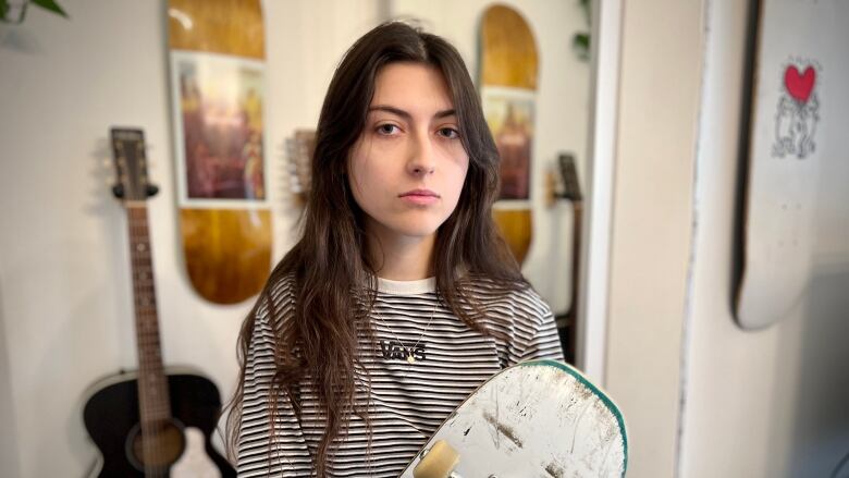 A woman with long dark hair wearing a black and white striped shirt holds a skateboard deck.