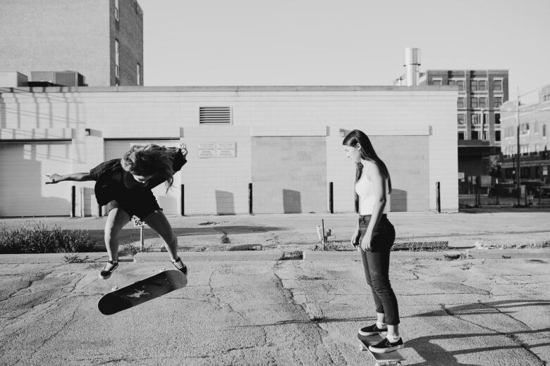 One person with long blonde hair does a kickflip on a skateboard, while another person with long dark hair stands on a skateboard watching.