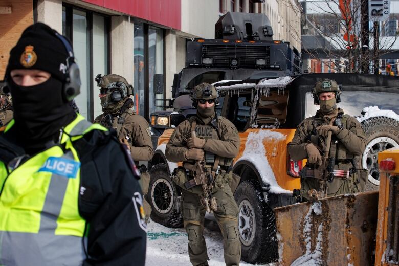 Police in green and brown tactical gear stand in a city on a winter day.