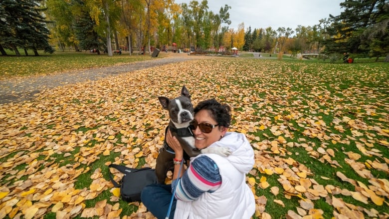 A woman holds a dog and sits on the grass surrounded by golden autumn leaves.