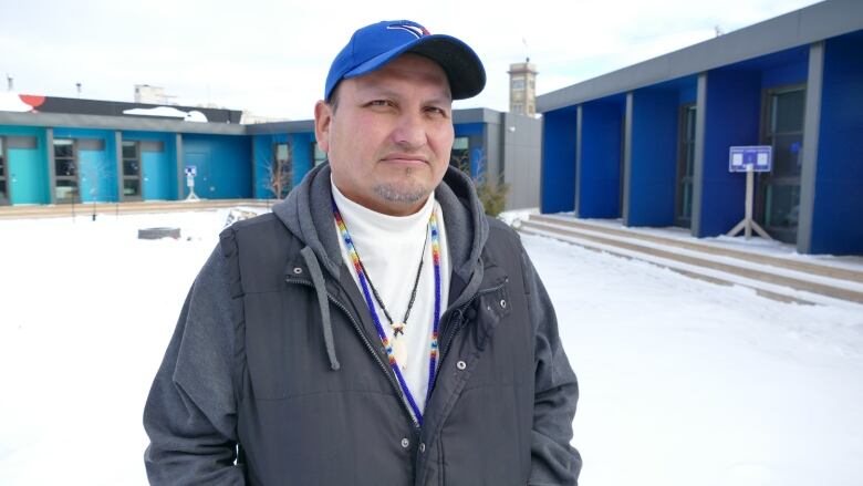 A man wearing a blue baseball cap and beaded necklace stands in an open courtyard, in front of rows of small houses.