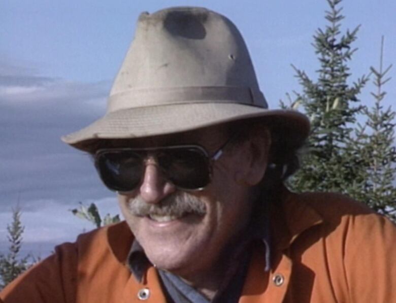 A man wears a brown hat, sunglasses and an orange jacket while smiling. He is outdoors. 