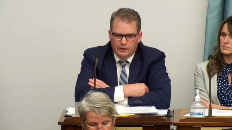 A man wearing a suit and tie with glasses and sitting at a desk speaks at the Prince Edward Island legislature. 