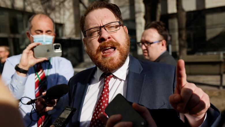 A lawyer in a suit gestures with a finger as he speaks, surrounded by reporters, outside a building.