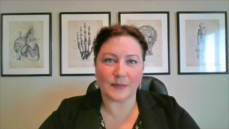 A woman with brown hair tied back sits against a white wall. There are four picture frames on the wall.