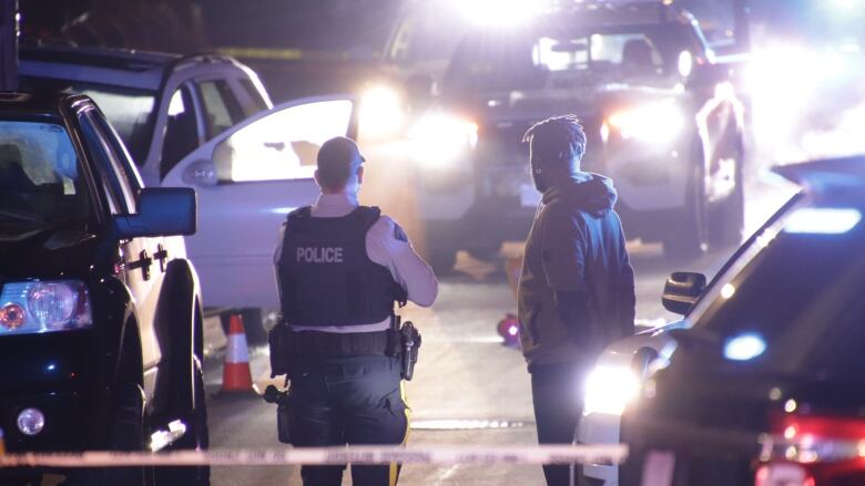 A police officer and a person in plainclothes stand with their backs to the camera in front of a number of vehicles with their lights on. Directly behind them is a white SUV with an open front door. The glass is shattered.