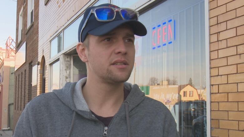 Man stands outside in a grey hoodie and ballcap. 