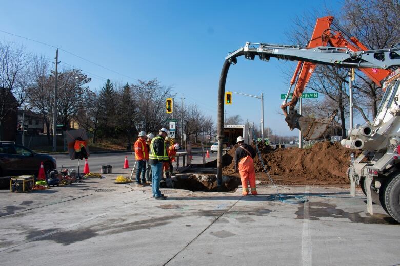 People wearing hard hats and safety vests stand around a hole in the road while a machine sticks a tube into it.