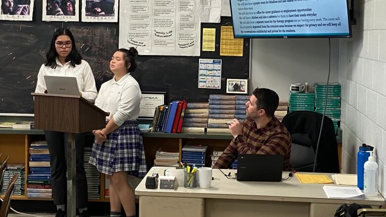 Two young students stand at a podium in front of a classroom speaking. Their teacher sits at his desk next to them watching. 