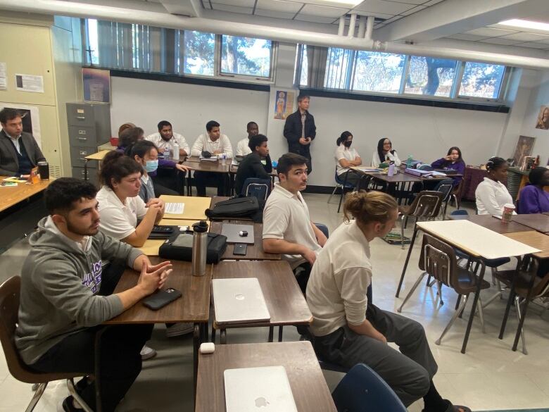 A classroom full of students listening to their peers give a presentation. A man sits in the back behind them.